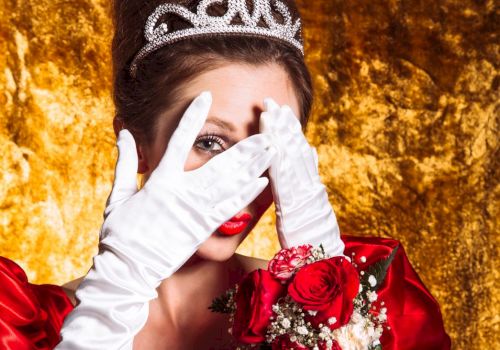 A woman wearing a tiara, white gloves, and holding roses covers her face with her hands against a gold background.