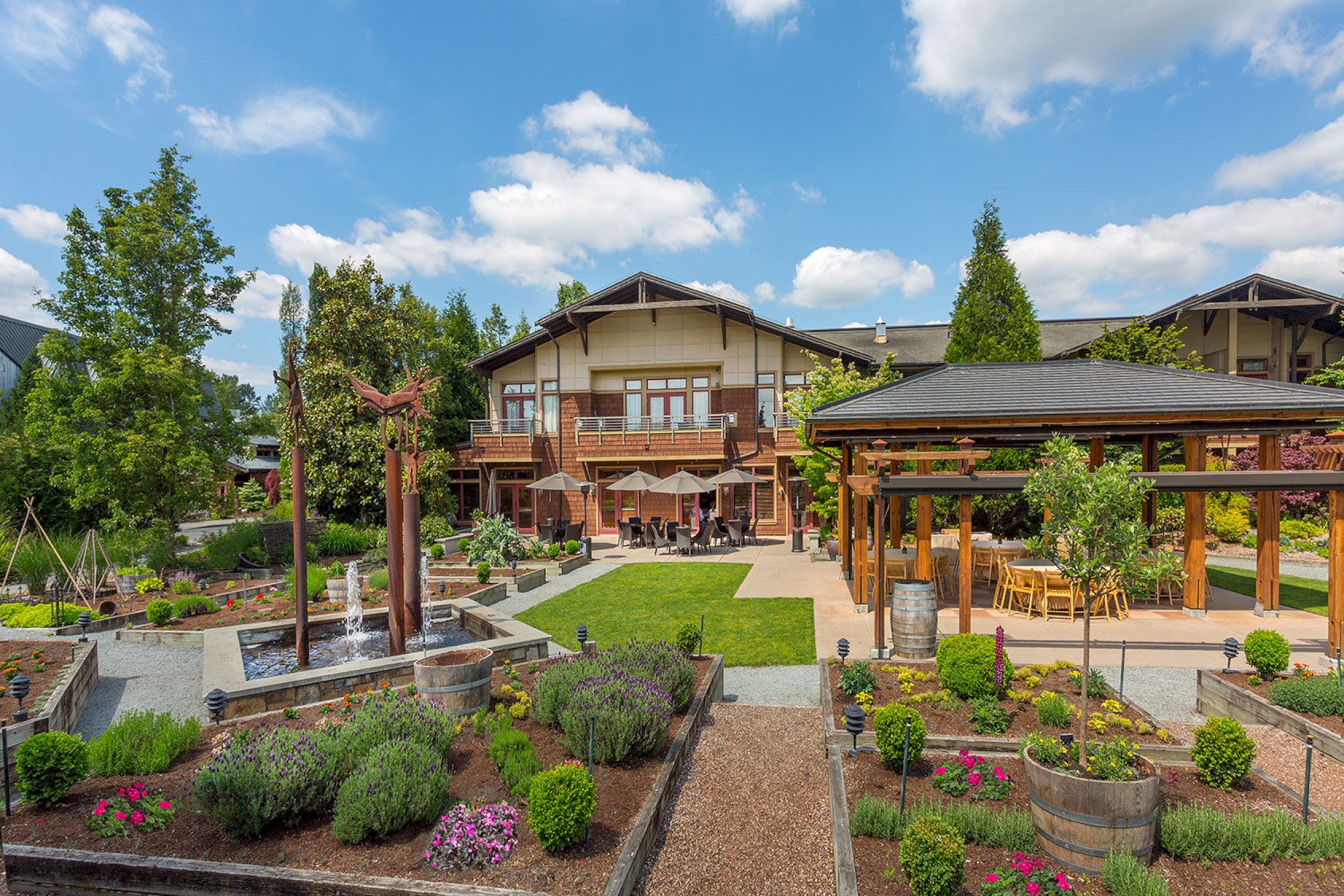 A large, well-maintained garden surrounds a two-story building with outdoor seating and greenery, under a bright, partly cloudy sky.