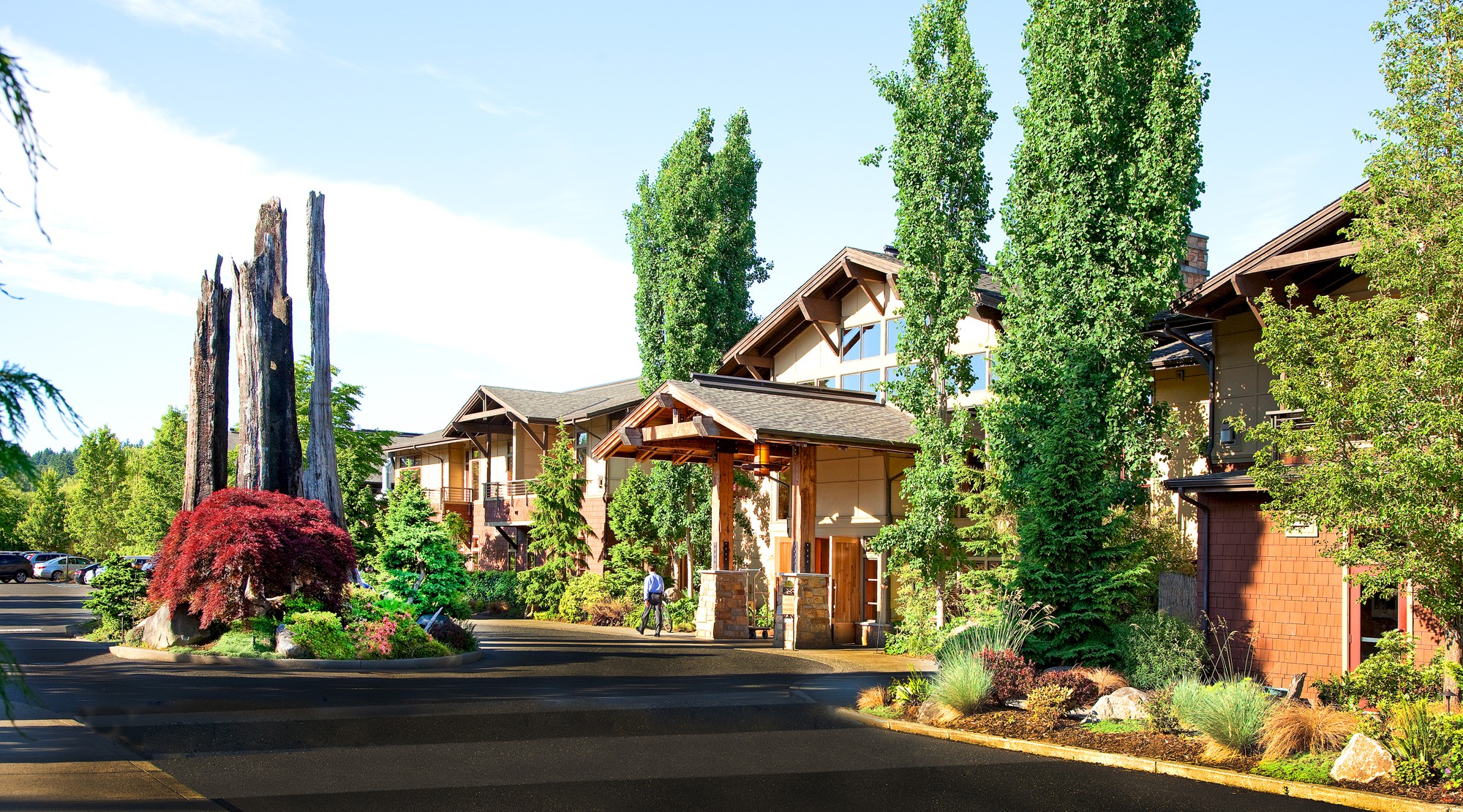 A picturesque lodge with tall green trees, a wooden entrance, and lush landscaping under a clear blue sky, showcasing a serene and inviting environment.