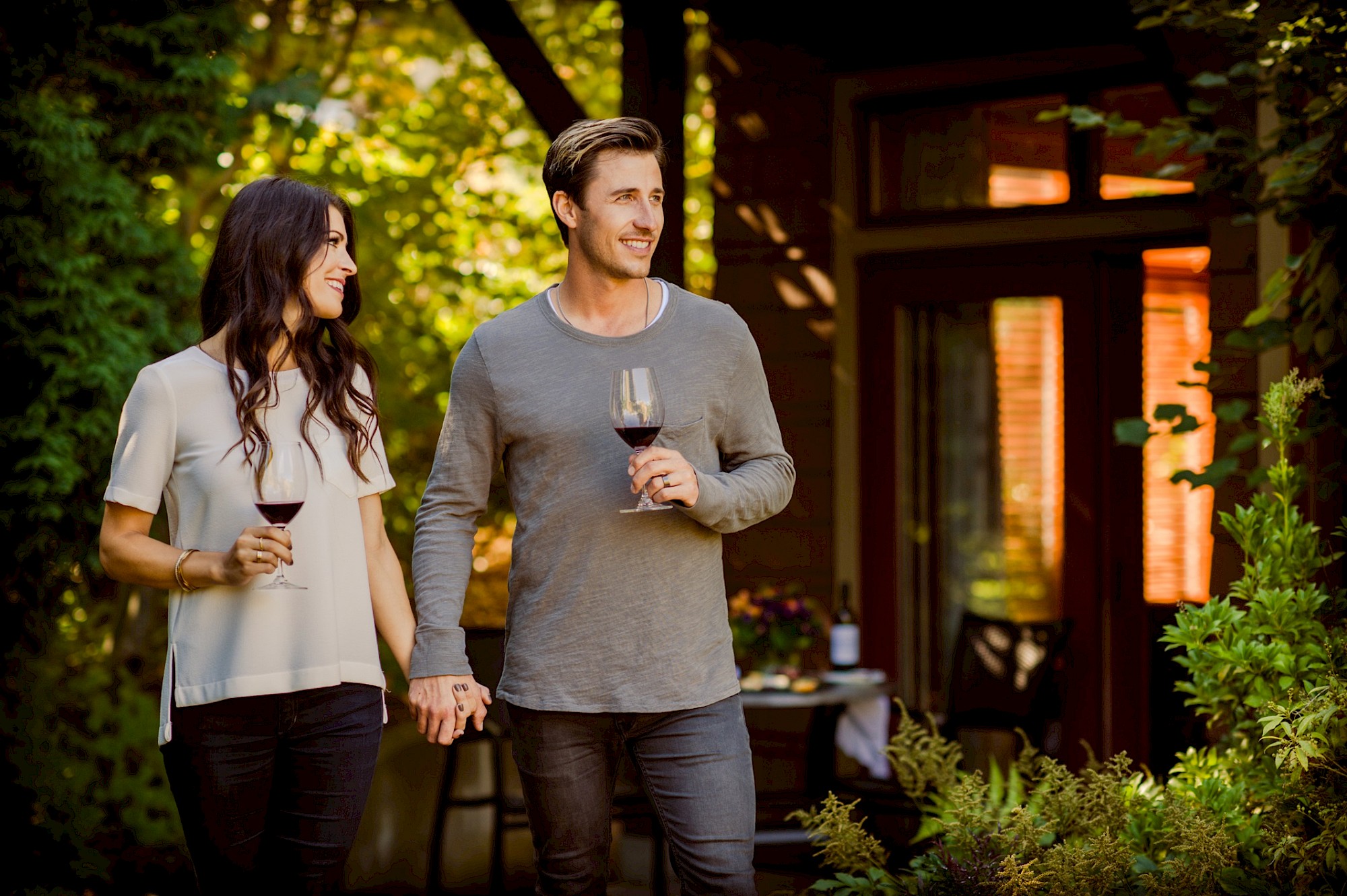 A couple walks hand in hand outdoors, each holding a glass of red wine, with a house and greenery in the background.