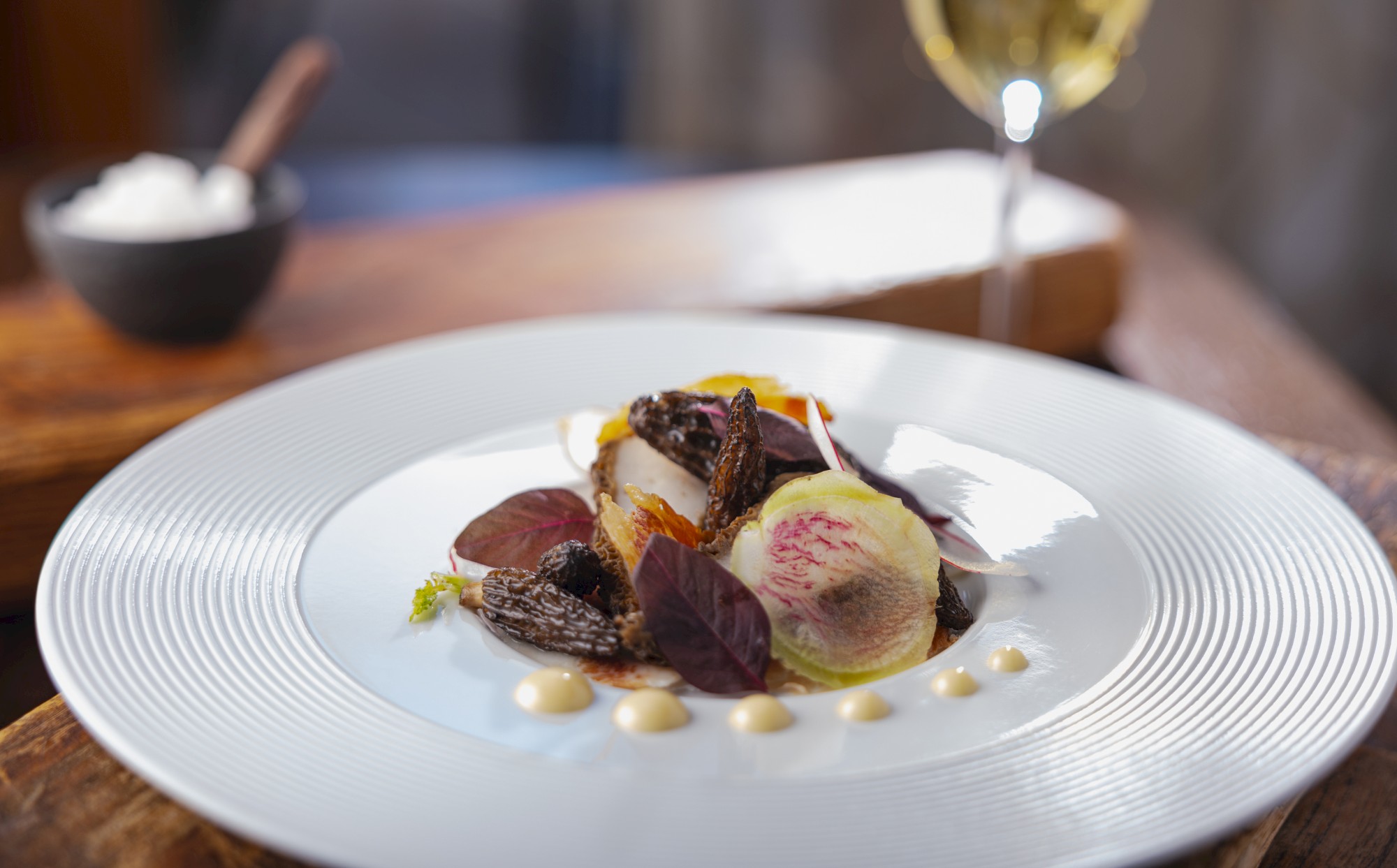 A gourmet dish featuring various vegetables and garnishes artistically plated on a white dish, accompanied by a glass of white wine.