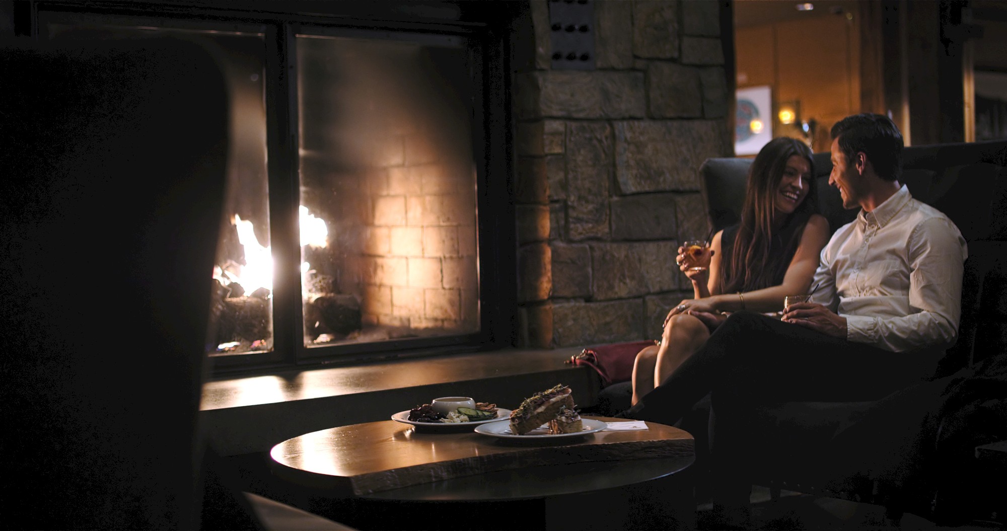 A couple is sitting by a fireplace, enjoying a cozy moment with drinks in hand, and a table with food in front of them.