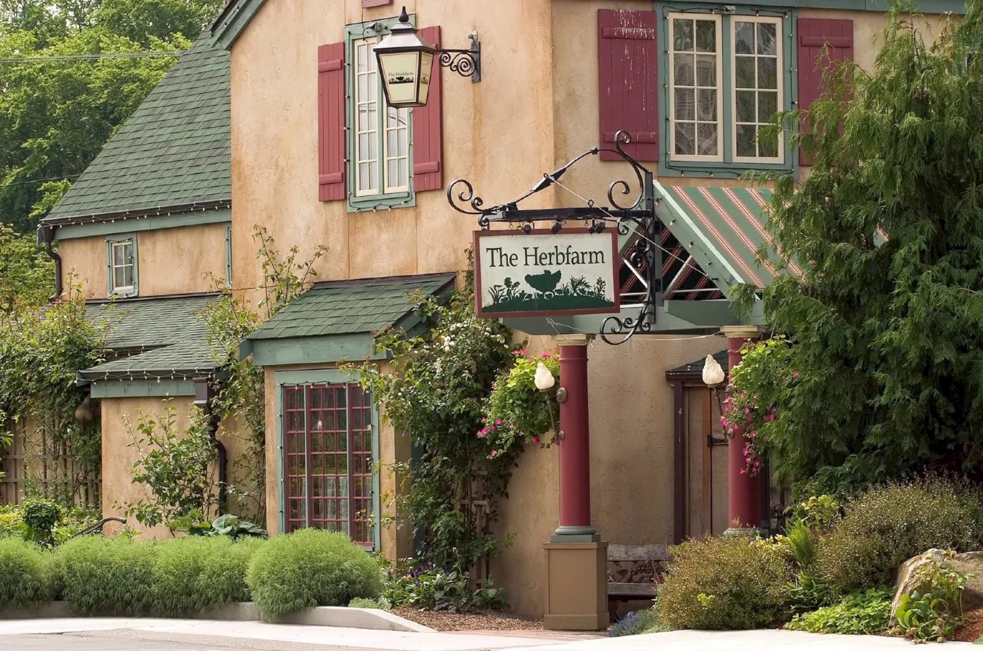 A charming restaurant or café with a rustic exterior, named The Herbfarm, featuring green and red shutters, surrounded by lush greenery and plants.