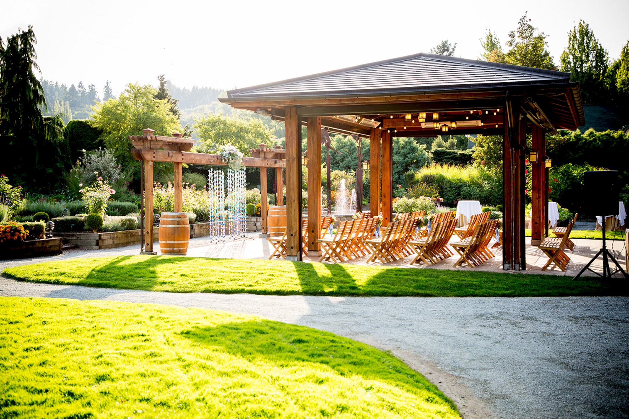 An outdoor pavilion with wooden chairs and well-kept greenery, set up for an event, under a sunny sky in a landscaped garden setting.