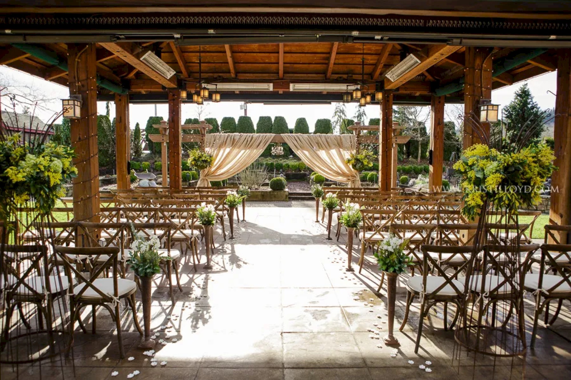 An outdoor wedding venue with wooden chairs, green floral decorations, and a draped fabric archway under a covered structure, arranged for a ceremony.