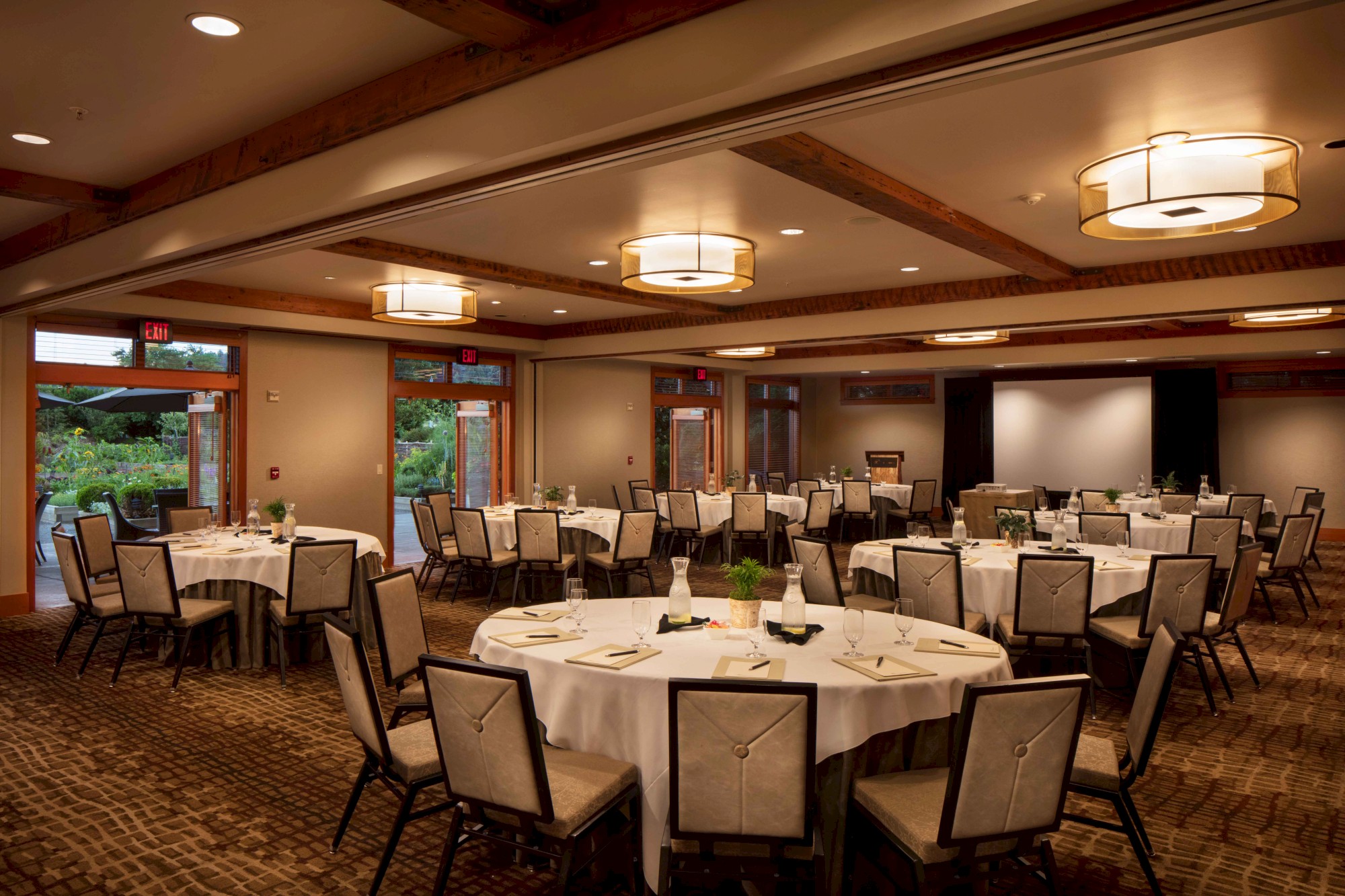 A large, elegant conference room is set up for an event with round tables covered in white cloths, neatly arranged chairs, and warm overhead lighting.