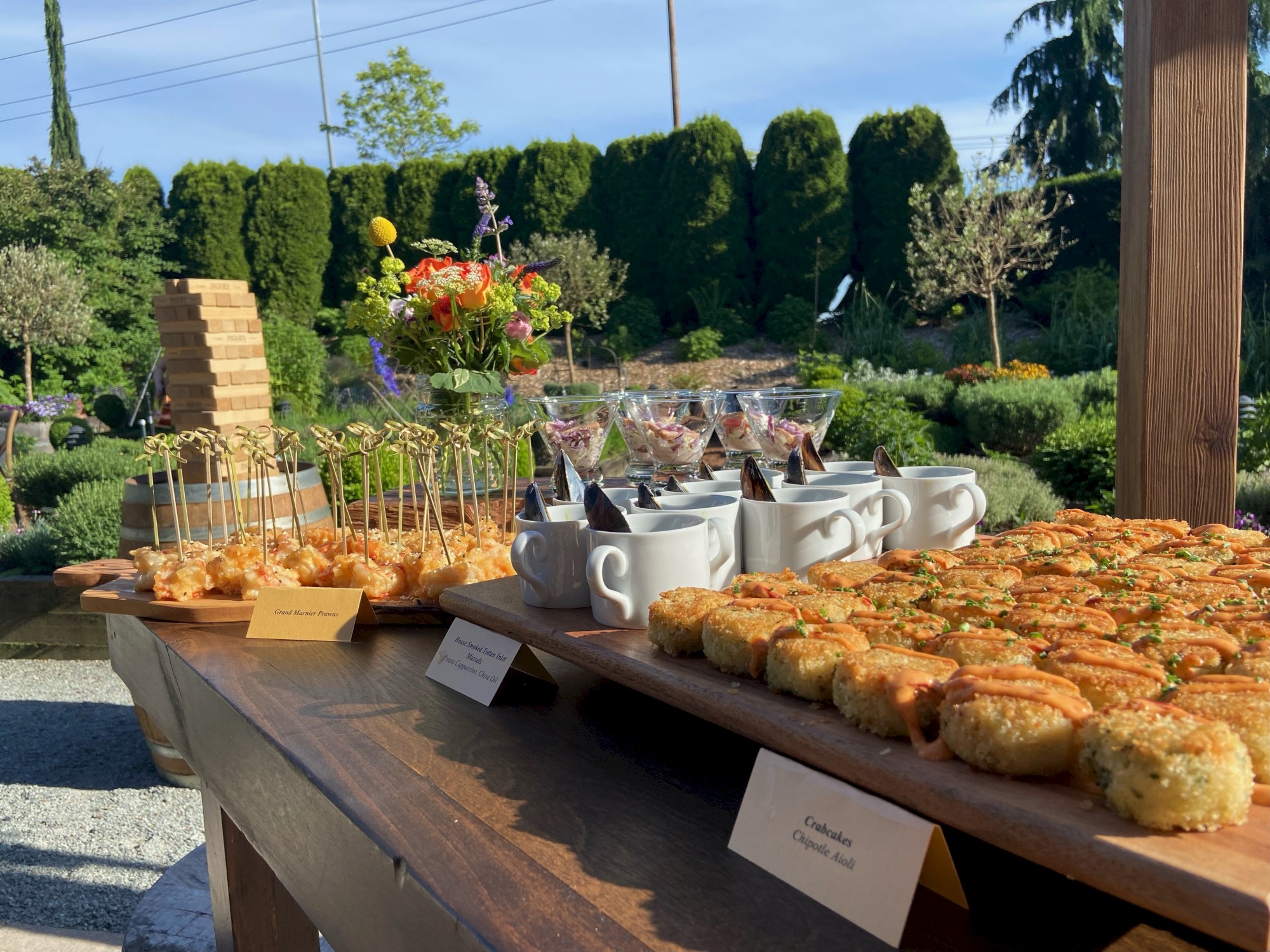 An outdoor buffet display with various appetizers, including cups of soup and fried bites, set against a garden backdrop with flowers and greenery.