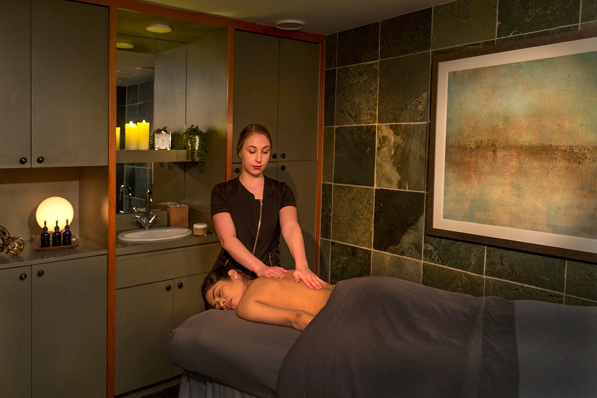 A massage therapist is giving a back massage to a client in a dimly lit, serene room with soft lighting and modern decor, including a large framed painting.