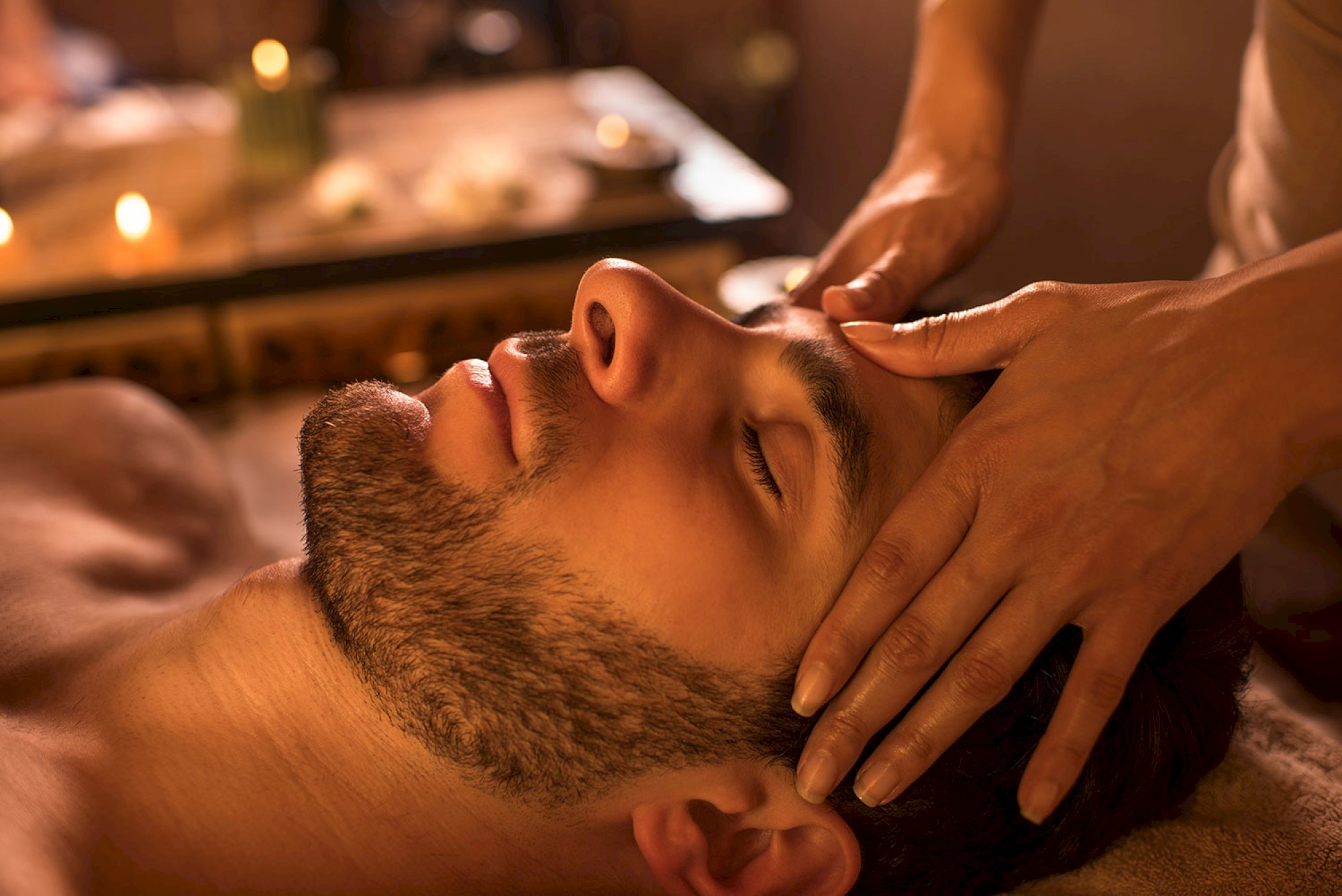 A person is receiving a relaxing head massage in a soothing, candle-lit environment, lying down with eyes closed and a peaceful expression.