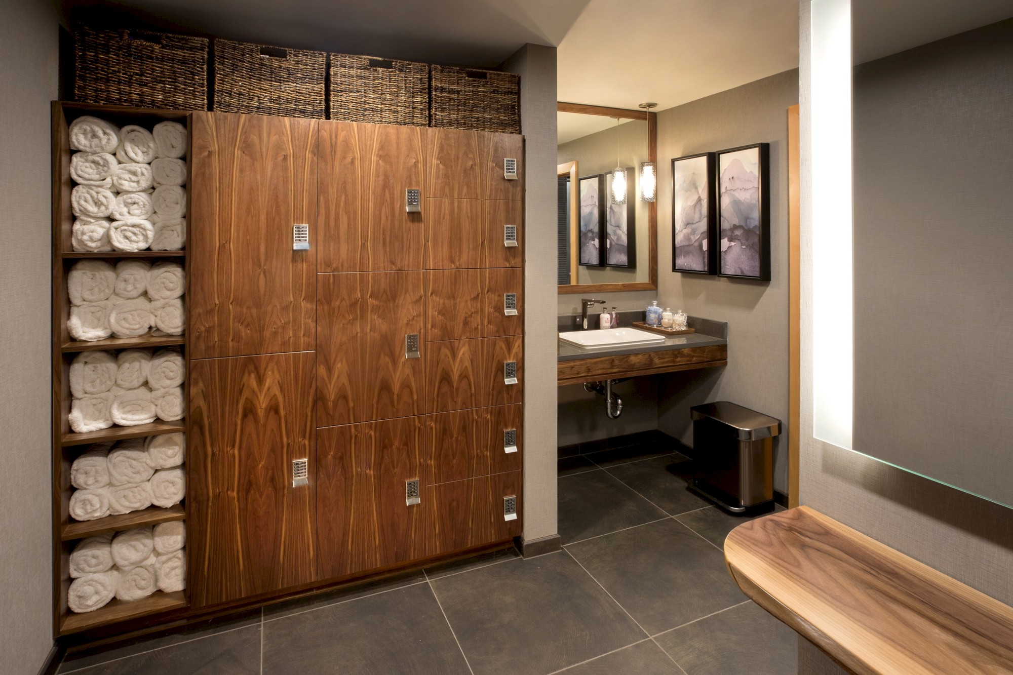 The image shows a locker room with wooden lockers and neatly stacked towels, a sink with a mirror, artwork on the wall, and a trash bin.