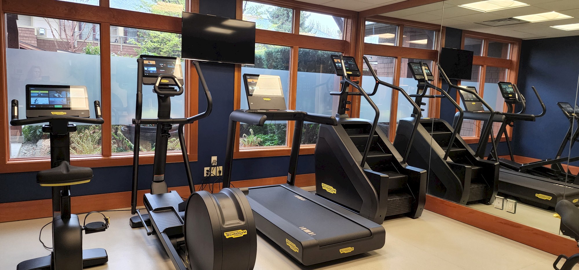 This image shows a gym with various exercise equipment, including treadmills, exercise bikes, and stair climbers, next to large windows and mirrors.