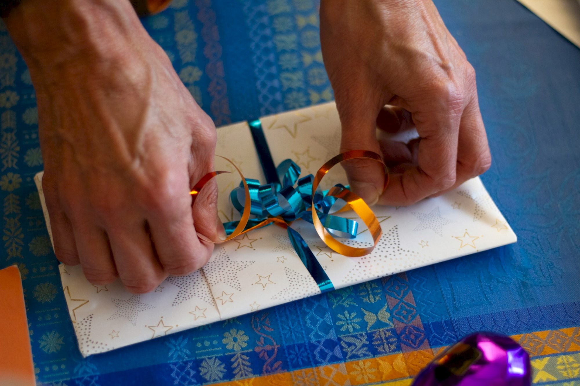 The image shows hands tying a blue and orange ribbon into a bow around a gift wrapped in white star-patterned paper.