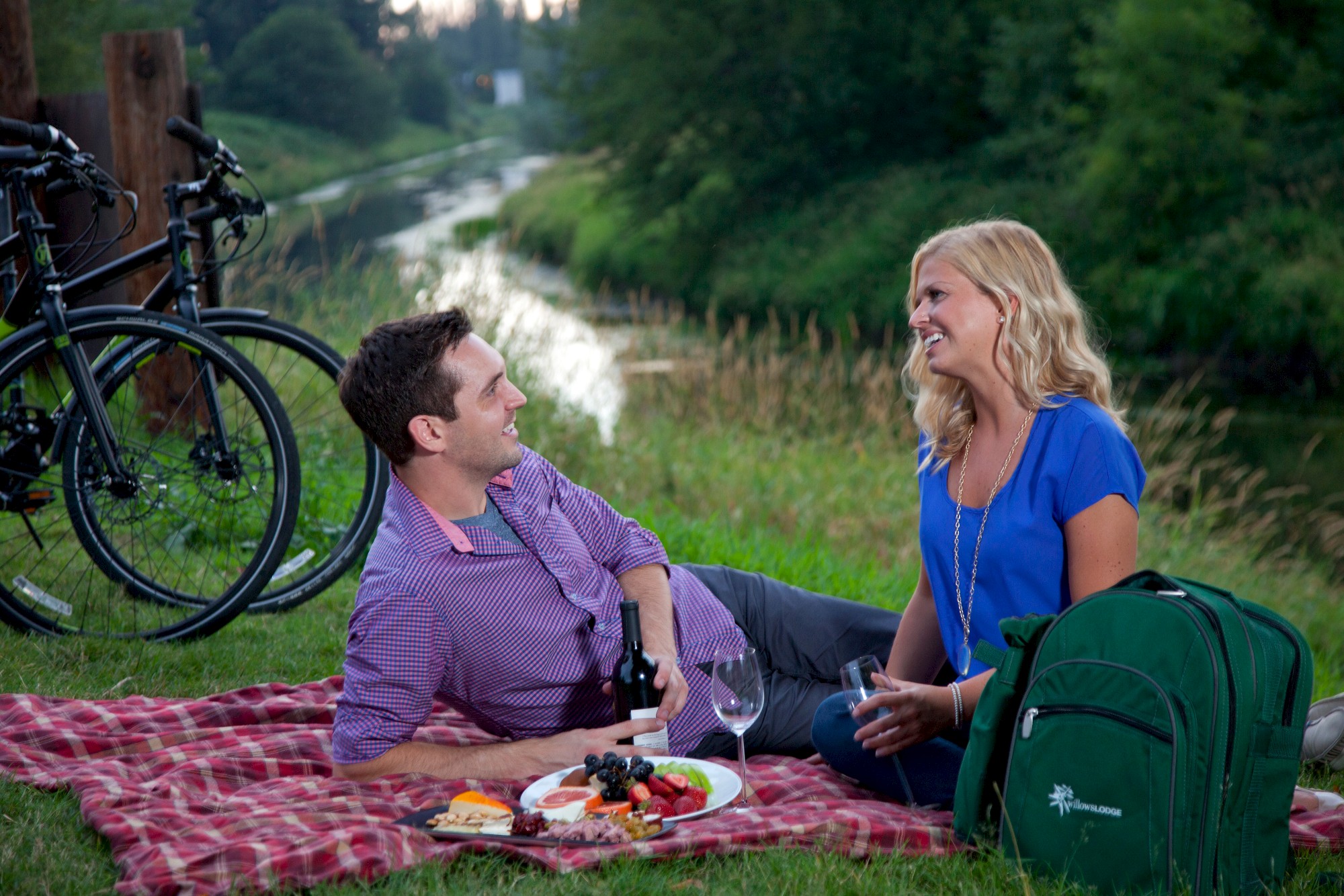 A couple enjoying a picnic by a river with bikes in the background. They're sitting on a red blanket with food, drinks, and a green backpack.