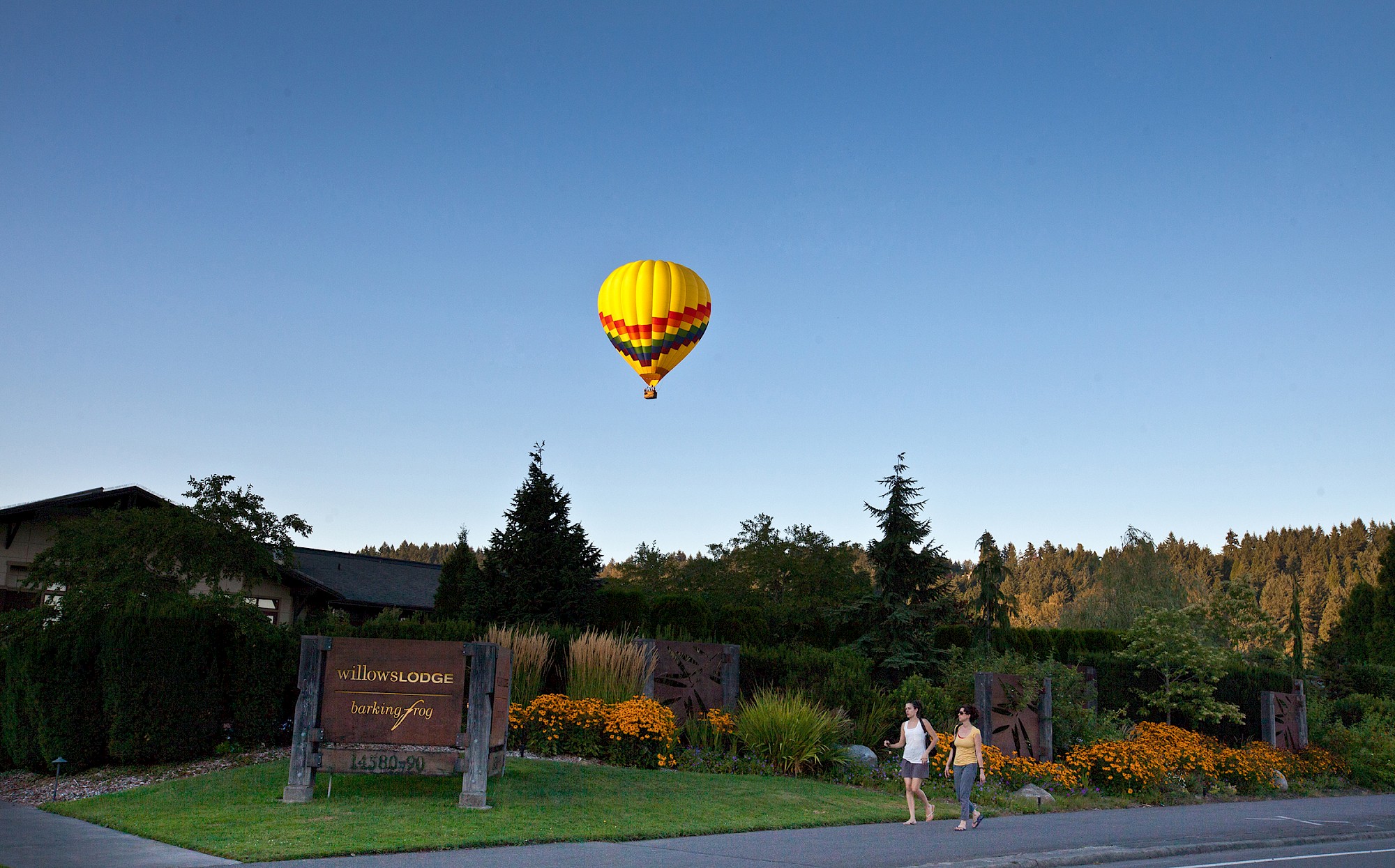 A hot air balloon flies over a scenic area with a building and gardens, a sign in front reads 