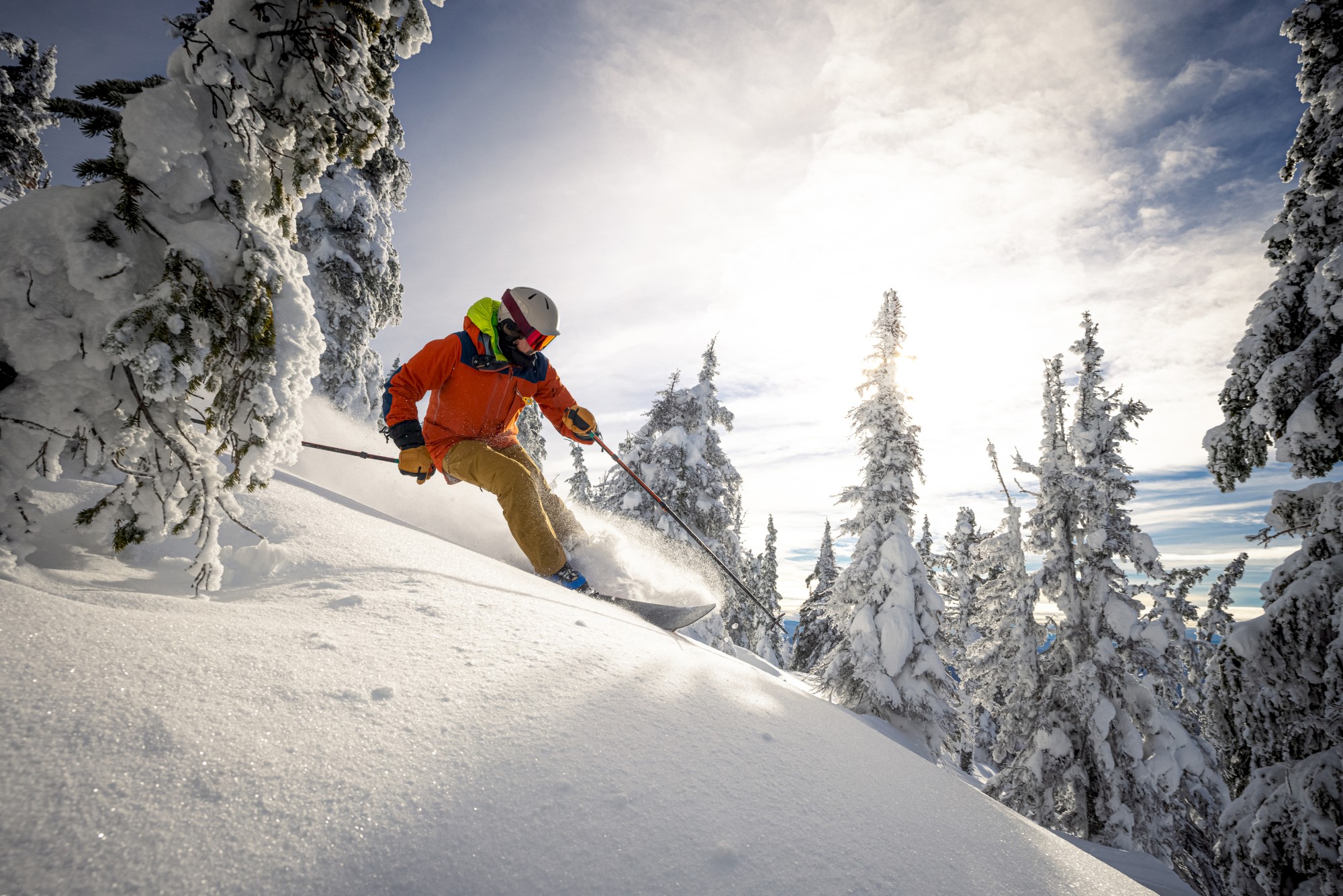 A skier in an orange jacket navigates through a snow-covered forest, surrounded by tall trees and a bright sky.