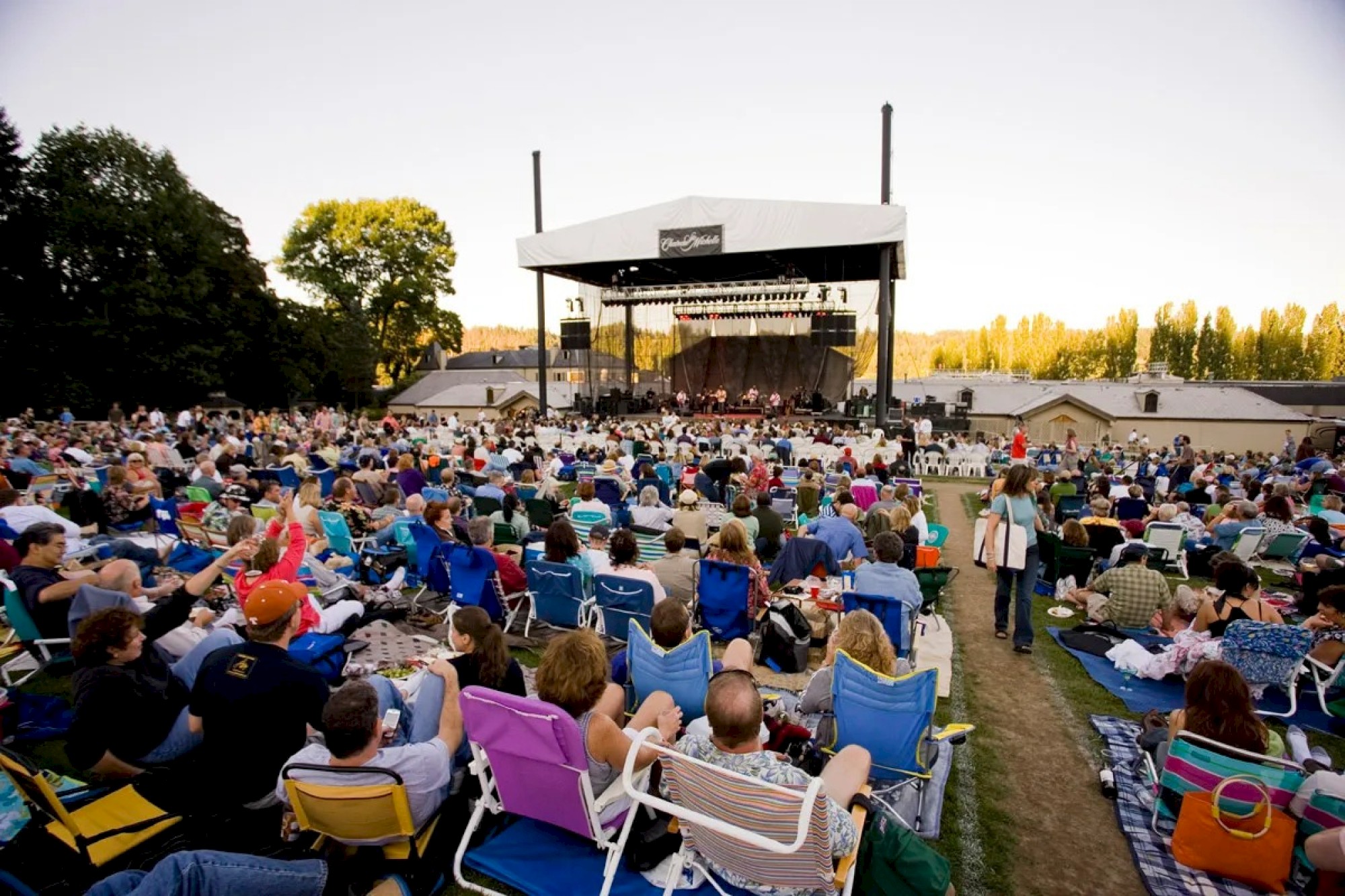 A large outdoor concert with many people seated on foldable chairs and blankets, facing a stage with performers under a white canopy.