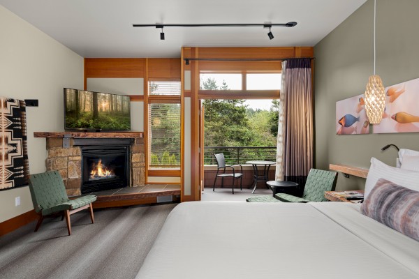 A cozy hotel room with a bed, fireplace, TV, chairs, and a balcony with a forest view, decorated with modern art and lighting.