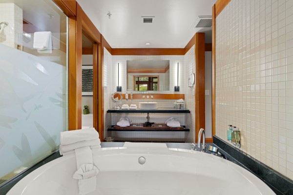 This image shows a bathroom with a large bathtub, towels, a mirror, and wooden frame decorations. A sink and toiletries are visible on the counter.