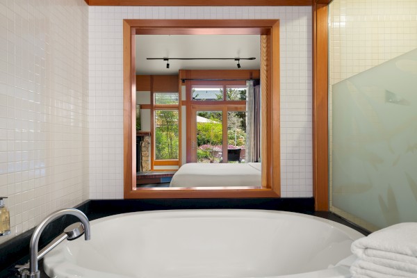 A bathtub with a rolled towel beside it faces a large window with a scenic outdoor view, framed by wooden accents.