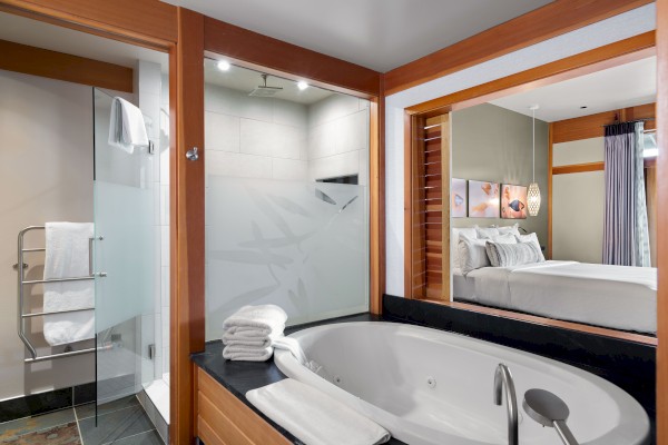 A modern bathroom featuring a bathtub, a folded towel, a glass-enclosed shower, and a view into an adjacent bedroom with a neatly made bed.