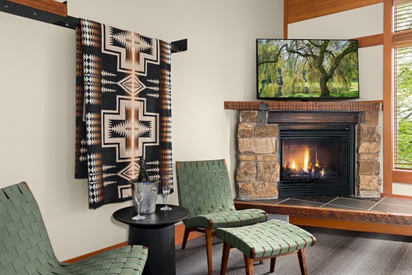 A cozy living room with a fireplace, two green woven chairs, a small table, a hanging patterned blanket, and a TV displaying a tree scene.