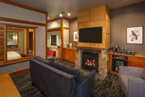 A cozy living area with a sectional sofa, fireplace, wall-mounted TV, adjacent kitchenette, and view of the bedroom through a window opening.