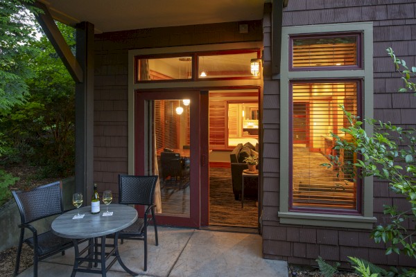 A cozy outdoor patio with a small table set for two, wine glasses, and chairs next to a windowed door leading into a warmly lit interior space.