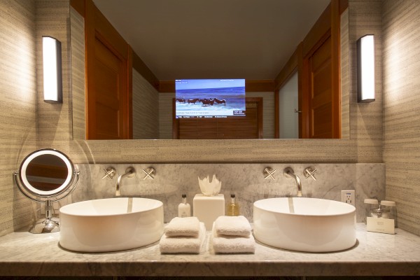 The image shows a modern bathroom with double sinks, a large mirror, a TV, towels, and toiletries neatly arranged on a marble countertop.