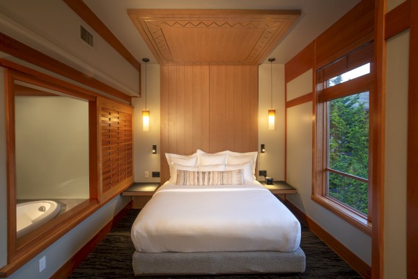 A cozy, modern bedroom with a neatly made bed, wooden accents, two side tables, wall lamps, large windows, and a view of greenery outside the windows.