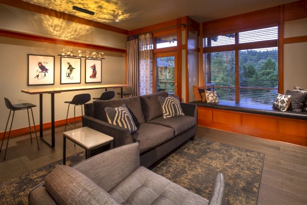 A modern living room with a gray sofa, armchair, high table with stools, framed bird artwork, and large windows with a scenic outside view.