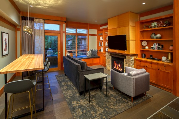 A cozy living room with a fireplace, TV, sofas, and a wooden dining area on the left. The room has warm lighting and large windows.