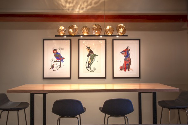 The image shows a modern dining area with a wooden table, four chairs, and three framed artworks of colorful, stylized animals on the wall.