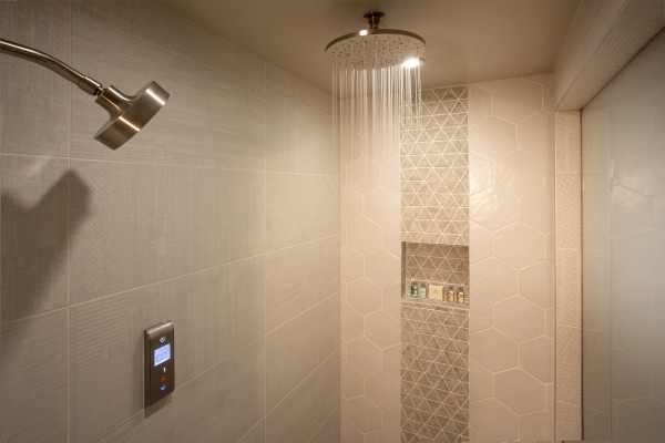 A modern shower with a rainfall showerhead, a handheld showerhead, geometric tile design, built-in shelf with toiletries, and smart controls.