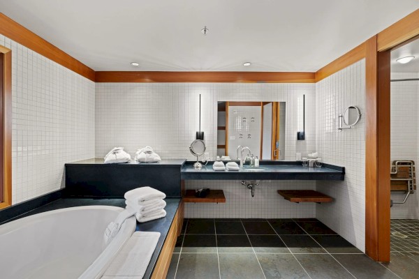 A modern bathroom featuring a bathtub, neatly folded towels, a large mirror with lights, and a spacious counter with amenities.