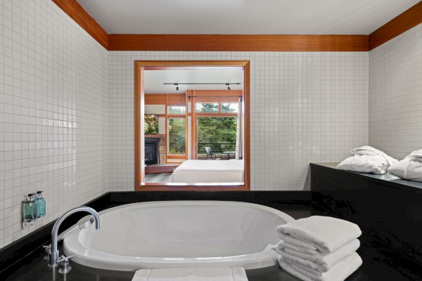 A bathroom with a large bathtub, folded towels, and a window view of a bedroom with large windows.