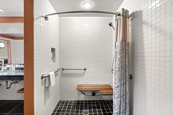 This image shows a bathroom with a walk-in shower featuring white tiles, a wooden bench, a shower curtain, and a grab bar. The sink is visible outside.