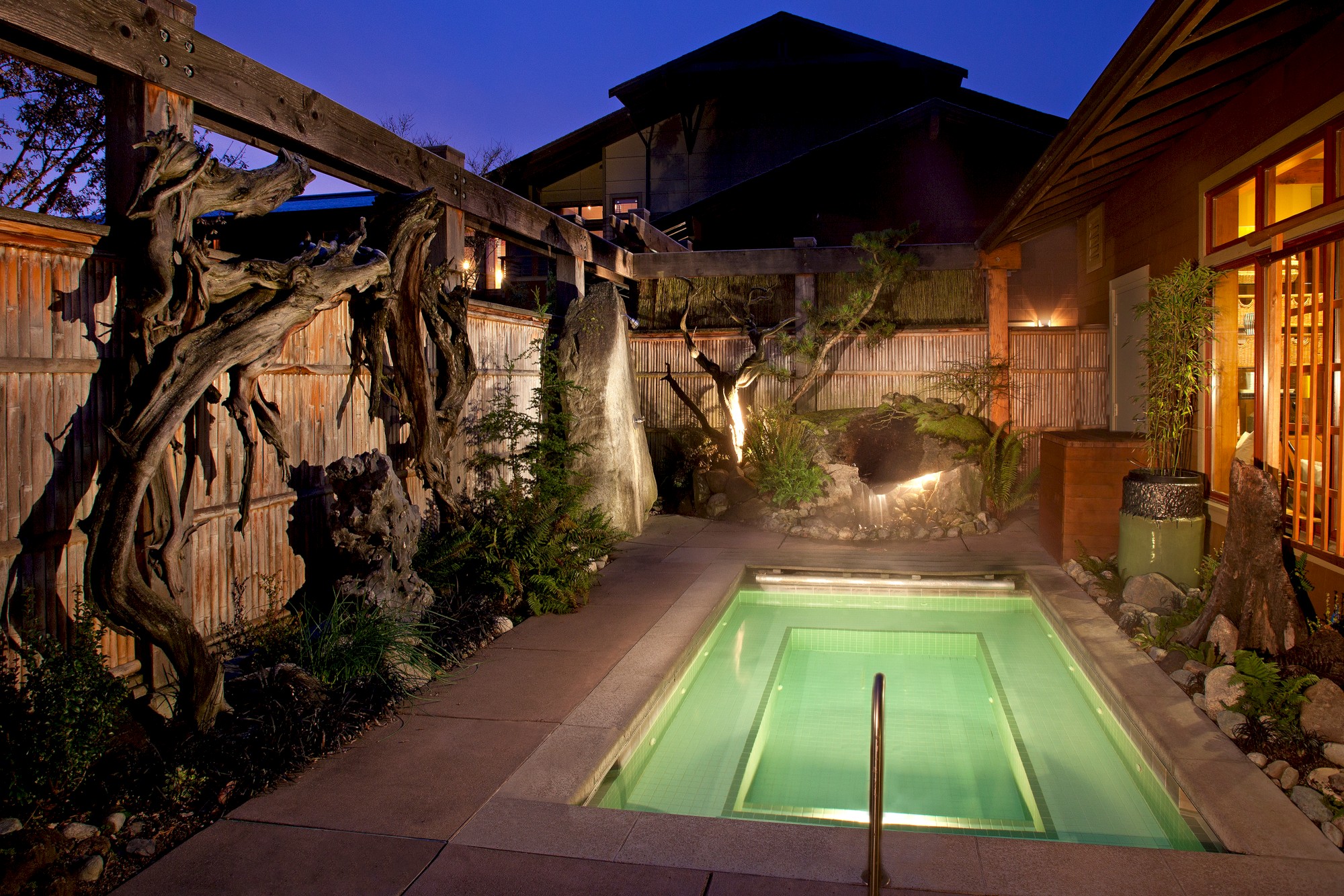 An illuminated outdoor hot tub is surrounded by a wooden fence, lush plants, and ambient lighting in a serene garden setting.