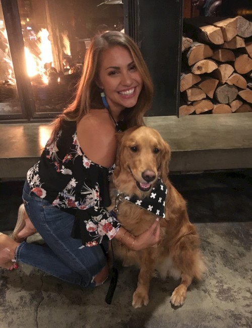 A woman is smiling and kneeling next to a golden retriever in front of a fireplace, with stacked firewood visible in the background.