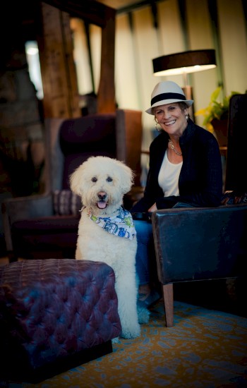 A person wearing a hat is sitting in a cozy room next to a large white fluffy dog with a scarf around its neck.