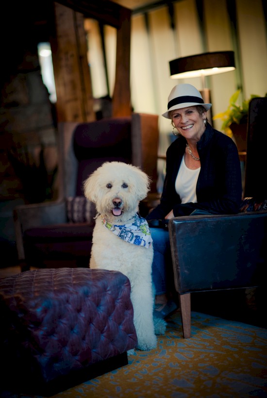 A person wearing a hat is sitting in a cozy room next to a large white fluffy dog with a scarf around its neck.