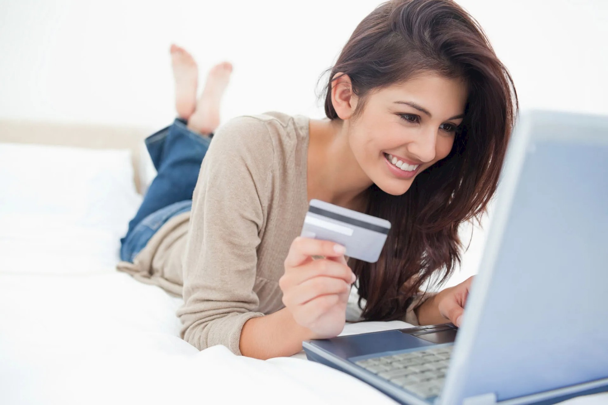 A woman is lying on her bed, smiling while holding a credit card and using a laptop for online shopping or browsing.