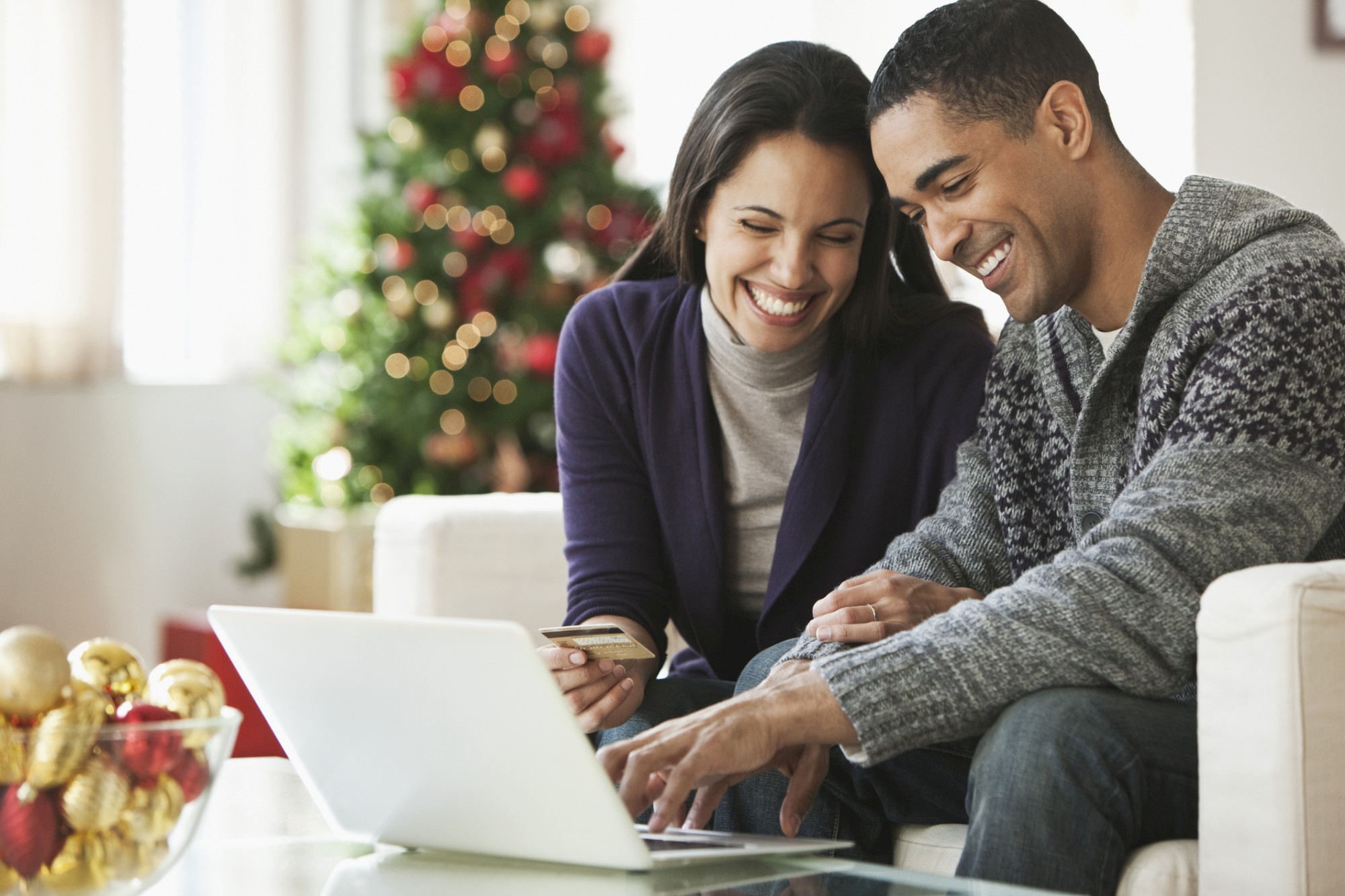 A couple sits on a sofa, smiling at a laptop screen, with a Christmas tree and decorations in the background.