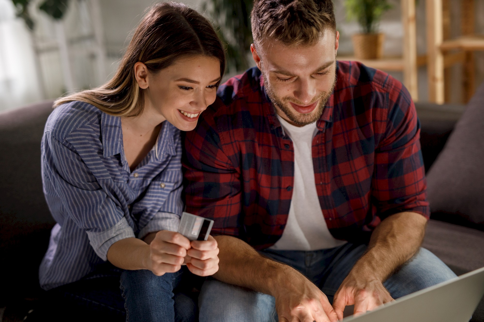 A couple is sitting on a couch, smiling while shopping online with a credit card and laptop in a cozy home setting.