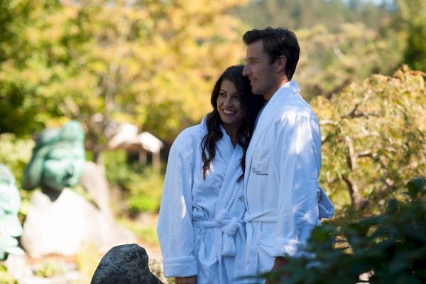 A couple in white robes is standing outside in a garden with greenery and sculptures in the background.