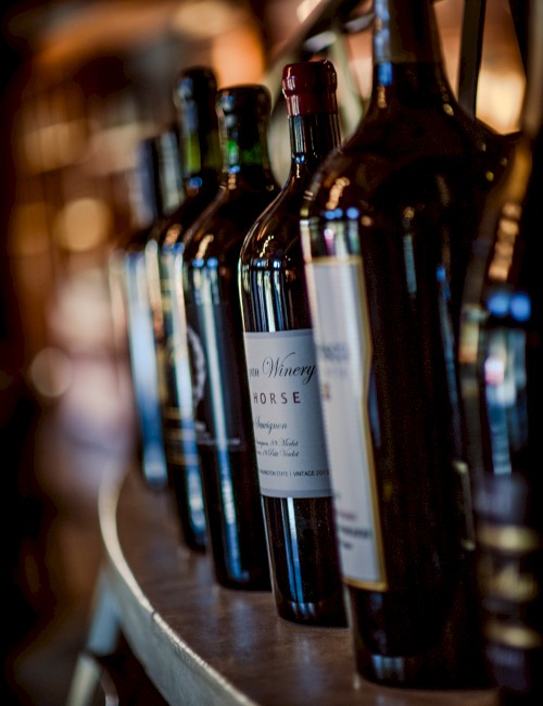 The image shows a row of wine bottles lined up on a wooden shelf, partially blurred, with labels indicating various wine brands.