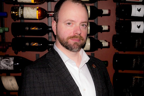 A man in a suit stands in front of a wine rack filled with various bottles. He appears to be in a wine cellar or a similar environment.