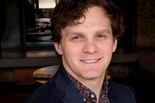 A person with curly hair is smiling, wearing a dark blazer over a patterned shirt, in an indoor setting.