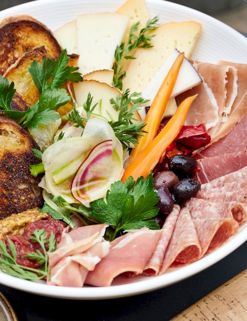 The image shows a charcuterie board with assorted meats, cheeses, toasted bread, pickled vegetables, olives, and fresh herbs, all arranged in a white bowl.