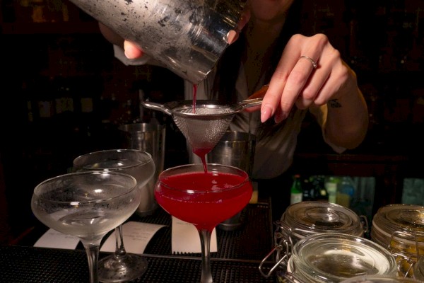 A person is pouring a red cocktail through a strainer into a glass at a bar. There are empty glasses and jars nearby.