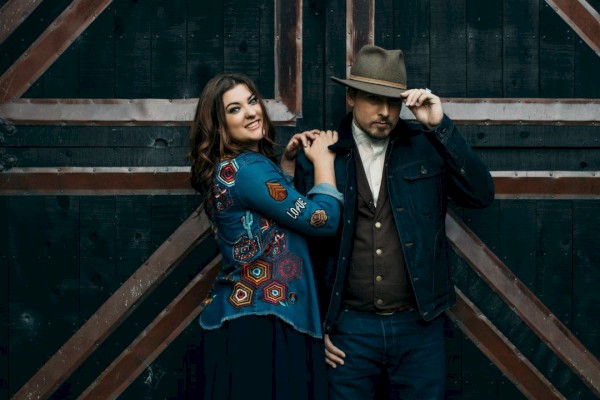 A man and woman stand in front of a rustic wooden door; the woman smiles, while the man holds his hat brim. Both are wearing stylish outfits.